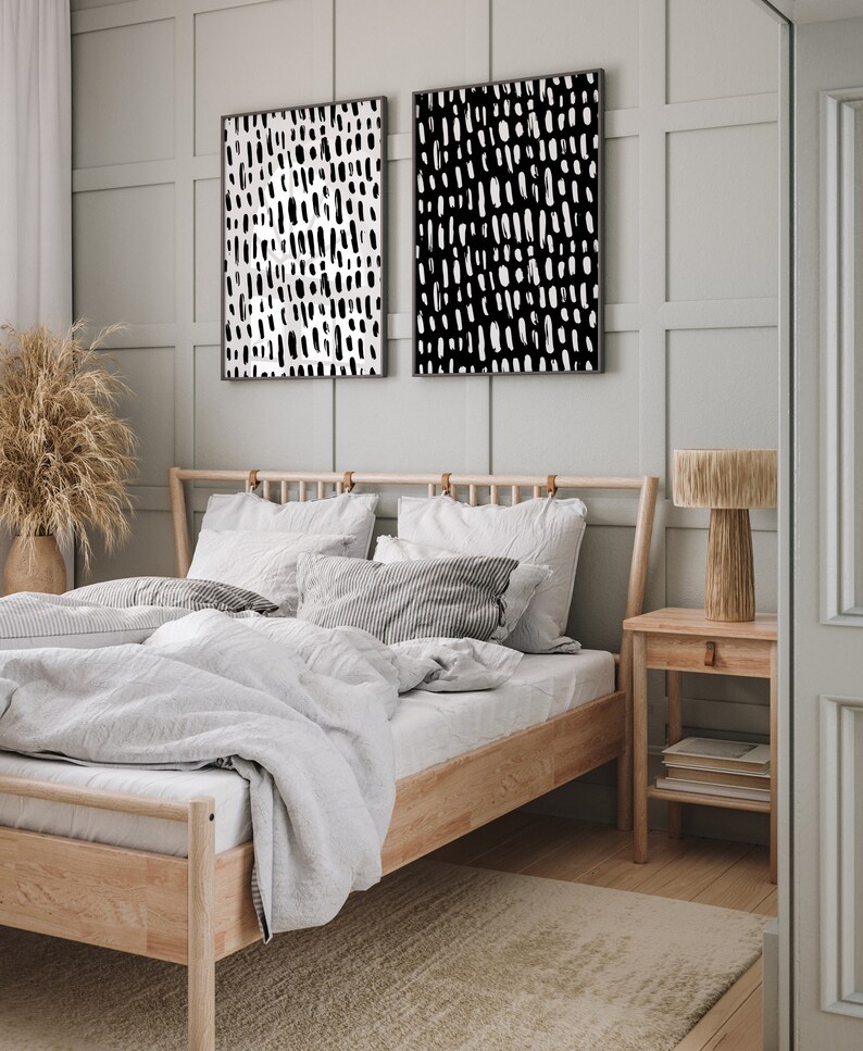 Puede incluir: Dos impresiones art&iacute;sticas abstractas enmarcadas con patrones de pinceladas en blanco y negro cuelgan sobre una cama con marco de madera clara y ropa de cama blanca. Una mesita de noche de madera con una l&aacute;mpara y libros se encuentra junto a la cama.