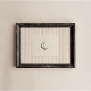 Op de afbeelding: Een rechthoekige donkerhouten lijst met een crèmekleurige passe-partout met dunne verticale zwarte strepen. Gecentreerd in de passe-partout bevindt zich een kleine, gebroken witte rechthoekige print met een zwarte inktillustratie van een maansikkel en drie kleine sterren.