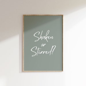 Op de afbeelding: Een ingelijste print met een saliegroene achtergrond en de witte tekst "Shaken or Stirred?". De print heeft een dunne, goudkleurige lijst en wordt tegen een witte muur getoond. De tekst is in cursief.