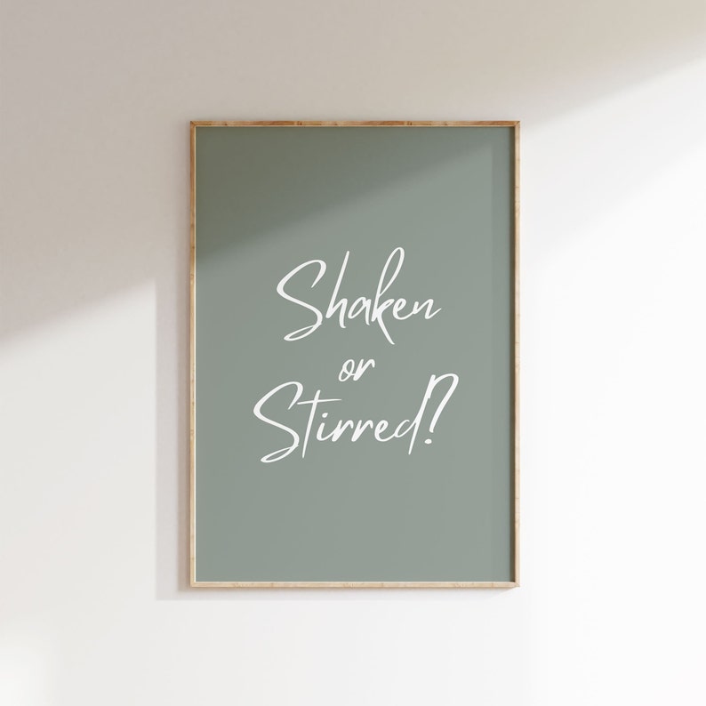 Op de afbeelding: Een ingelijste print met een saliegroene achtergrond en de witte tekst "Shaken or Stirred?". De print heeft een dunne, goudkleurige lijst en wordt tegen een witte muur getoond. De tekst is in cursief.