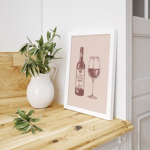 May include: A framed print featuring a wine bottle and a wine glass, both filled with red wine. The artwork is in a sketch style with the words "Red Wine" on the bottle. A white pitcher with green leaves sits to the left.