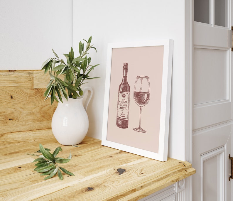 May include: A framed print featuring a wine bottle and a wine glass, both filled with red wine. The artwork is in a sketch style with the words "Red Wine" on the bottle. A white pitcher with green leaves sits to the left.