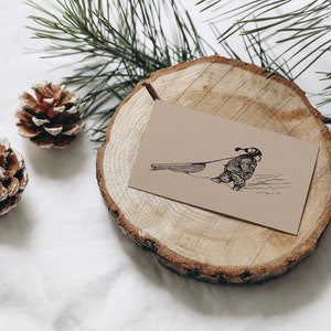 May include: A tan card with a black illustration of a person pulling a sled in the snow rests on a wooden slice. Two pine cones and pine branches are also present, creating a winter theme.