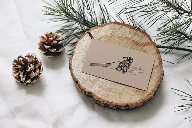 May include: A tan card with a black illustration of a person pulling a sled in the snow rests on a wooden slice. Two pine cones and pine branches are also present, creating a winter theme.