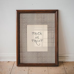 May include: A rectangular wall art piece in a dark brown wooden frame featuring a cream-coloured centre panel with the black text "TRICK OR TREAT" written in a stylised script font, surrounded by a vertical black and white pinstripe border.