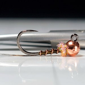 May include: A small, copper-colored bead fly fishing lure with a brown and gold body and a pink and yellow tail.