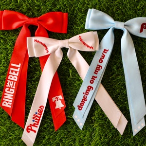 May include: Three hair bows with long tails. The bows are red, white, and light blue. The tails are decorated with the words "Ring the Bell", "Phillies", "Dancing on my own", and a baseball graphic.