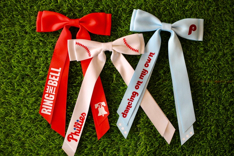 May include: Three hair bows with long tails. The bows are red, white, and light blue. The tails are decorated with the words "Ring the Bell", "Phillies", "Dancing on my own", and a baseball graphic.