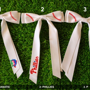 May include: Three white baseball bow ties with red stitching. The bow ties have different designs: one with a cartoon mascot, one with the word "Phillies", and one with a red "P".