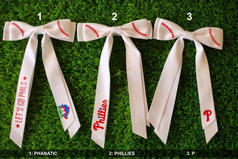 May include: Three white baseball bow ties with red stitching. The bow ties have different designs: one with a cartoon mascot, one with the word "Phillies", and one with a red "P".