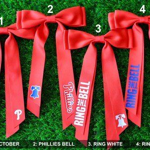May include: Four red ribbon bows with different designs. The first bow has the text "RED OCTOBER" printed on it. The second bow has the text "Phillies" and a Liberty Bell graphic. The third bow has the text "RING THE BELL" in white letters. The fourth bow has the text "RING THE BELL" in blue letters.