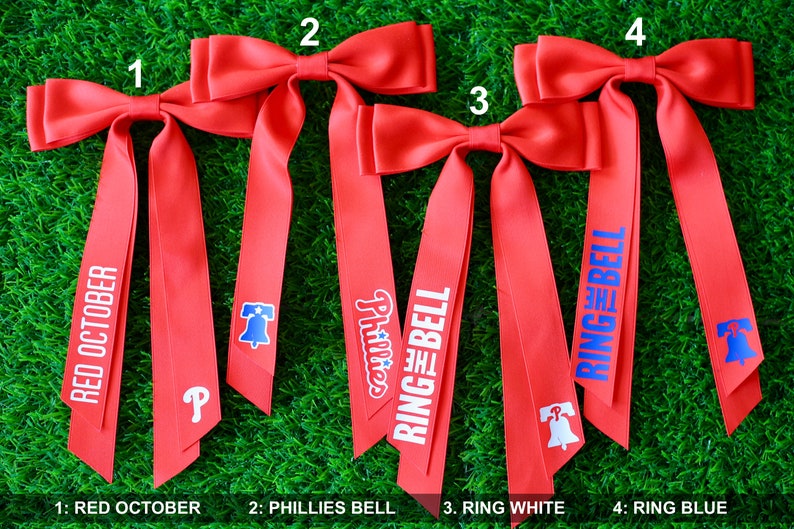 May include: Four red ribbon bows with different designs. The first bow has the text "RED OCTOBER" printed on it. The second bow has the text "Phillies" and a Liberty Bell graphic. The third bow has the text "RING THE BELL" in white letters. The fourth bow has the text "RING THE BELL" in blue letters.