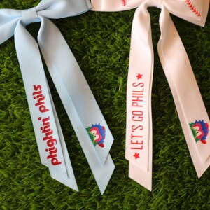 May include: Two blue and white ribbon hair bows with baseball-themed designs. One bow has the text "Fightin' Phils" and the other has the text "Let's Go Phils".
