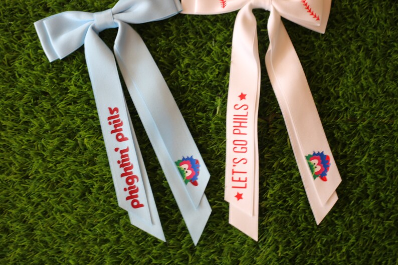 May include: Two blue and white ribbon hair bows with baseball-themed designs. One bow has the text "Fightin' Phils" and the other has the text "Let's Go Phils".