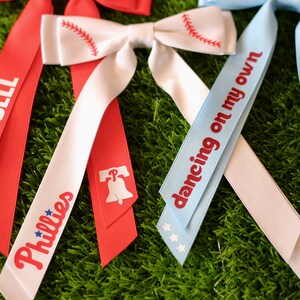 May include: Three red, white, and blue ribbon bows with different text on each. The first bow says "Ring the Bell" on the red ribbon and "Phillies" on the white ribbon. The second bow has a baseball graphic on the white ribbon and a Liberty Bell graphic on the red ribbon. The third bow says "dancing on my own" on the blue ribbon.
