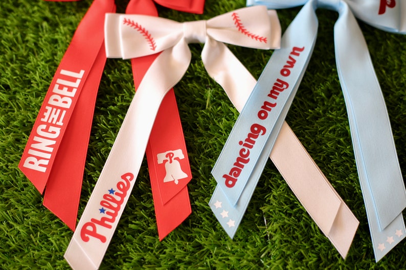 May include: Three red, white, and blue ribbon bows with different text on each. The first bow says "Ring the Bell" on the red ribbon and "Phillies" on the white ribbon. The second bow has a baseball graphic on the white ribbon and a Liberty Bell graphic on the red ribbon. The third bow says "dancing on my own" on the blue ribbon.