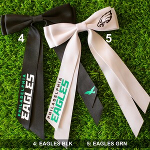 May include: Two black and white bow ties with the text "Philadelphia Eagles" printed on them. The bow tie on the left is black with white text and the bow tie on the right is white with black text. The bow tie on the right also has a small green eagle logo on the bow.