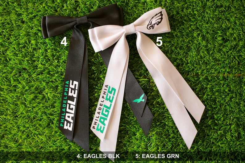 May include: Two black and white bow ties with the text "Philadelphia Eagles" printed on them. The bow tie on the left is black with white text and the bow tie on the right is white with black text. The bow tie on the right also has a small green eagle logo on the bow.