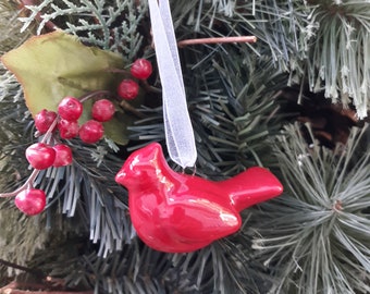 May include: Red ceramic cardinal Christmas ornament with a white ribbon hanging from a green artificial Christmas tree.