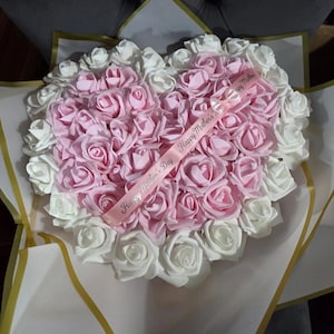 May include: A heart-shaped bouquet of artificial roses featuring a dense centre of light pink flowers surrounded by a border of white roses. A light pink ribbon with the printed text Happy Mother's Day is draped diagonally across the flowers.