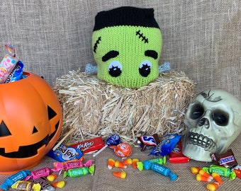 May include: A green and black knitted monster hat with stitched-on eyes and a pile of sweets, including Kit Kat, Snickers, and assorted fruit sweets, in front of a pumpkin and a skull.