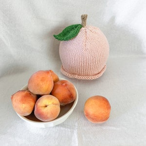 Pode incluir: Uma tigela de pêssegos com um chapéu de pêssego de malha. Os pêssegos são de cor laranja claro e o chapéu é de cor rosa claro.