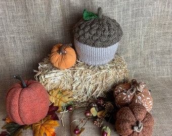 Puede incluir: Un gorro de bellota de punto marrón y beige con una hoja verde en la parte superior, rodeado de tres calabazas decorativas sobre un fondo de arpillera.