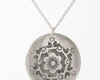 May include: A silver-toned pendant necklace with a circular design. The pendant features a central flower surrounded by a floral pattern. The necklace has a delicate chain and is displayed against a white background.