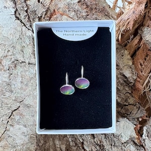 May include: A pair of oval-shaped earrings with silver-toned metal settings and iridescent, multi-colored surfaces featuring green and purple hues. The earrings are displayed in a white box with a black insert labeled The Northern Light Hand made.