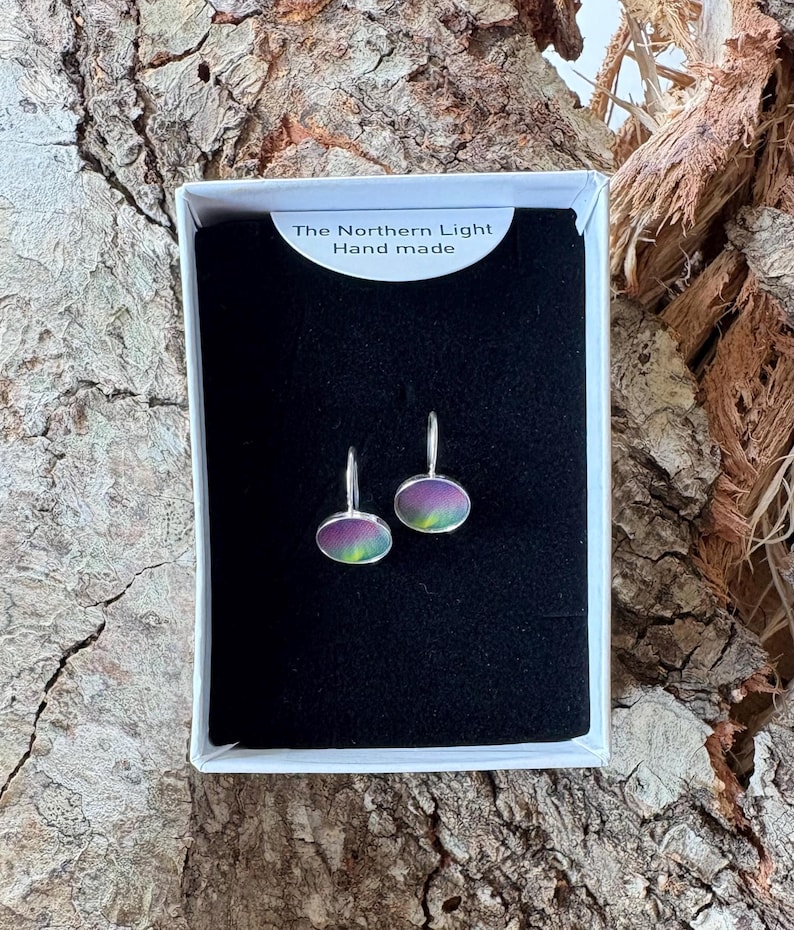 May include: A pair of oval-shaped earrings with silver-toned metal settings and iridescent, multi-colored surfaces featuring green and purple hues. The earrings are displayed in a white box with a black insert labeled The Northern Light Hand made.