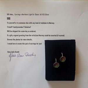 May include: A pair of 10mm AG 925 silver earrings labeled Northern Light B, featuring circular discs with a dark, iridescent green and purple surface, displayed on a black rectangular card with printed text and a signature by Hans Arne Buodd.