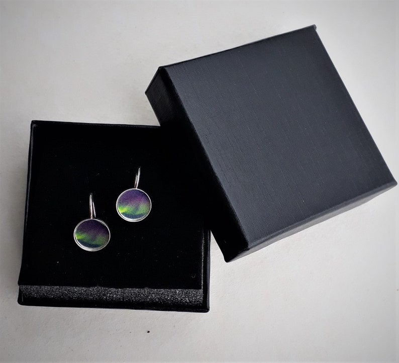 May include: A pair of round drop earrings featuring a vibrant purple and green aurora borealis pattern set in silver-toned metal frames, presented inside a square black jewelry box with a black foam insert and a matching black lid.