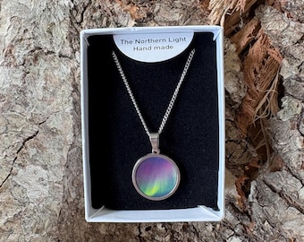 May include: A silver-toned circular pendant necklace featuring a colorful aurora borealis design in shades of purple, blue, and green. The necklace is presented in a white box with a black insert, labeled with the text The Northern Light Hand made.