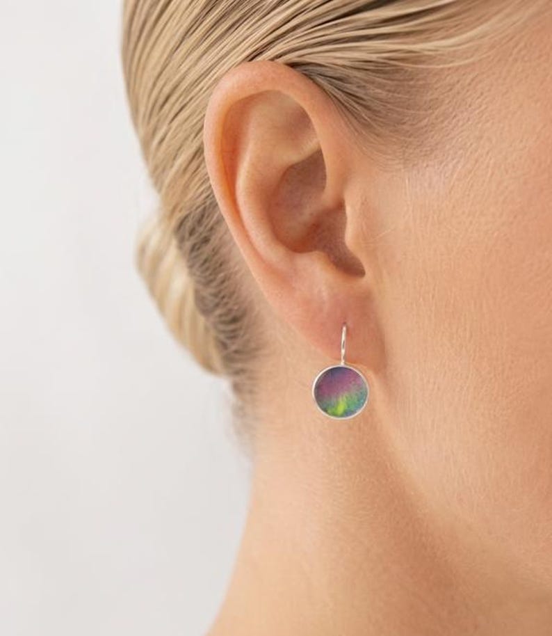 May include: A circular drop earring featuring a metallic silver frame and a vibrant, multi-colored inlay with swirling hues of purple, pink, and green. The earring hangs from a simple silver hook, displaying a smooth, polished surface finish.