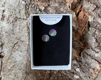 May include: A pair of circular stud earrings featuring a vibrant purple and green aurora borealis pattern. The earrings are displayed in a white rectangular box with a black insert, labeled with the text The Northern Light Hand made.