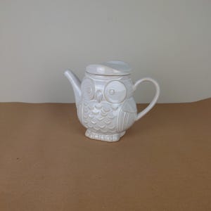 Puede incluir: Tetera de cerámica blanca con forma de búho y tapa. La tetera tiene un diseño de búho detallado con ojos grandes y plumas.