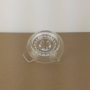 May include: A clear glass citrus juicer with a textured surface and a small spout in the center. The juicer has two handles on the sides.