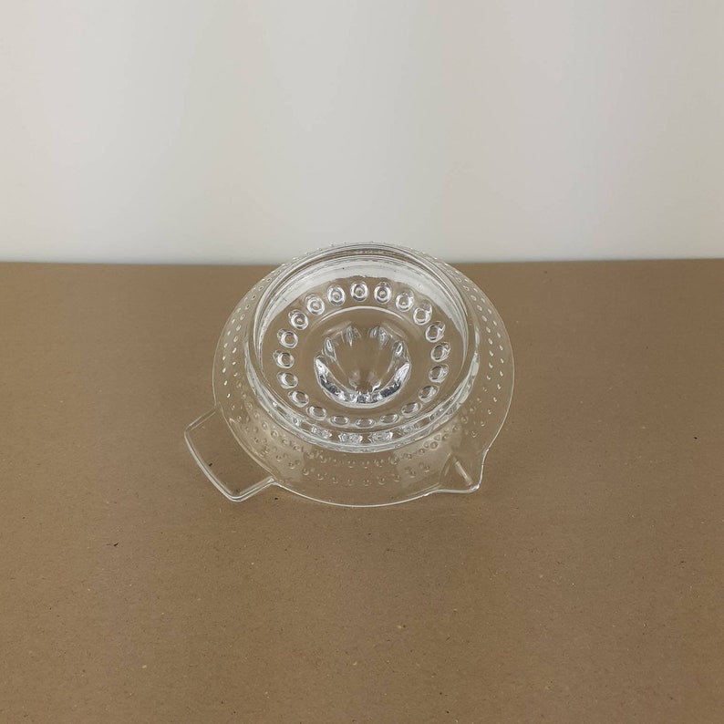 May include: A clear glass citrus juicer with a textured surface and a small spout in the center. The juicer has two handles on the sides.