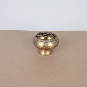 May include: A gold metal incense burner with an intricate design and a wire mesh top. The burner has a round base and is sitting on a brown surface.