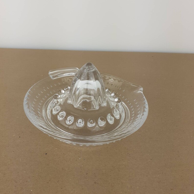 May include: A clear glass citrus juicer with a small, pointed cone and a wide, flat base with a textured rim. The juicer has a handle on the side.