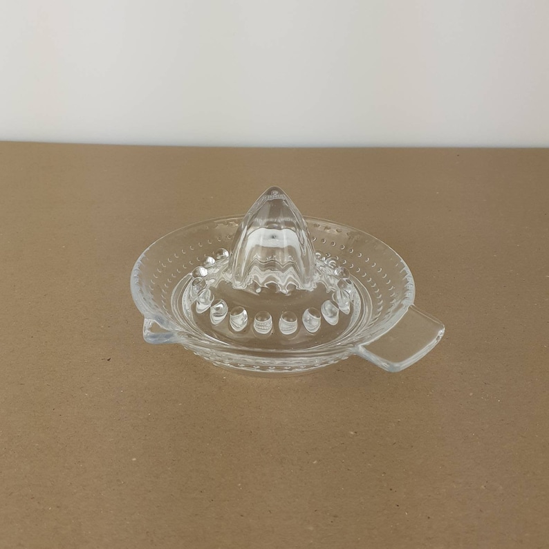 May include: A clear glass citrus juicer with a pointed top and a wide base. The base has a textured design and a handle on each side.