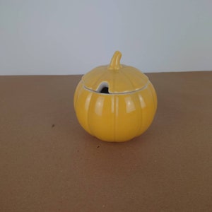 Op de afbeelding: Een gele keramische suikerpot in de vorm van een pompoen met een deksel. Het deksel heeft een klein gaatje om te gieten.