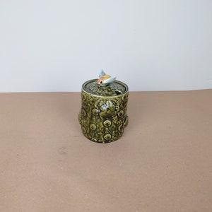 Op de afbeelding: Een groene keramische honingpot in de vorm van een boomstronk met een bij op de deksel. De pot heeft een bloemenpatroon en een klein gaatje om honing uit te gieten.