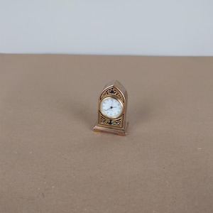 May include: A small gold-toned clock with black accents and an ornate design. The clock face is white with black numbers and hands.