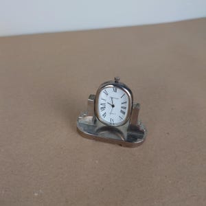 May include: Silver desk clock with Roman numeral markings on a white face. The clock is on a silver stand with a rectangular base.