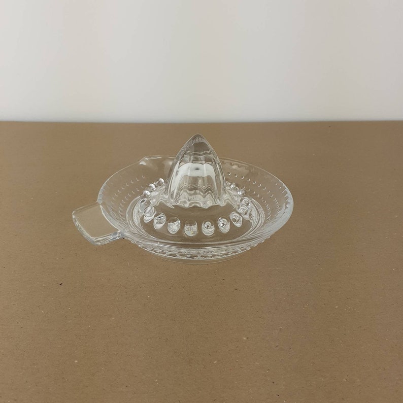 May include: A clear glass citrus juicer with a pointed top and a wide base. The base has a textured rim and a small handle.