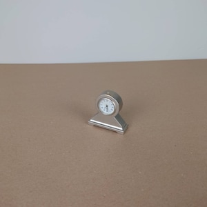 May include: A silver-toned tabletop clock with a white face and black hands. The clock is on a small stand with a rectangular base.