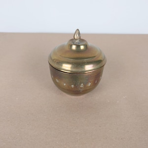 Op de afbeelding: Een klein, messing, dekselvormig bakje met een decoratief ontwerp. Het deksel heeft een puntige knop.