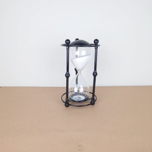 May include: A black metal hourglass with a clear glass bulb filled with white sand. The hourglass is on a brown surface.