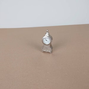 May include: A miniature silver-toned clock with a white face and black hands. The clock is shaped like a traditional grandfather clock.